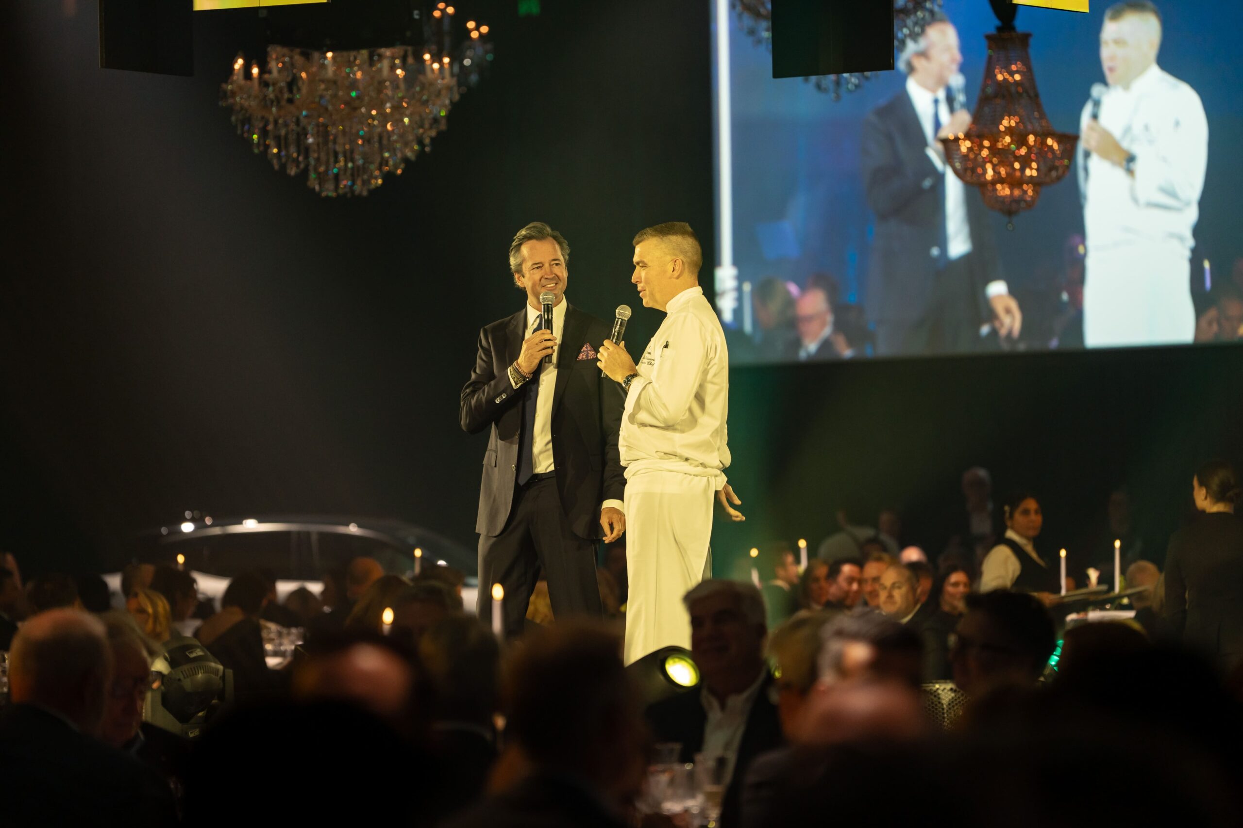 Two men stand on stage holding microphones and talking to each other at the formal Million Dollar Lunch 2025 event, with a seated audience and chandeliers in the background; their image is displayed on a large screen behind them.