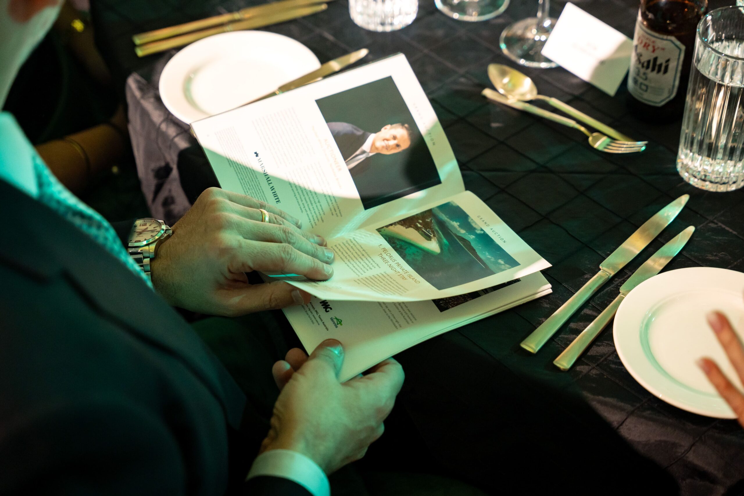 A person in formal attire sits at a table set for a meal at Million Dollar Lunch, holding and reading a booklet featuring a photo of a man and an image of a landscape or island. Plates, cutlery, and drinks are arranged on the black tablecloth.