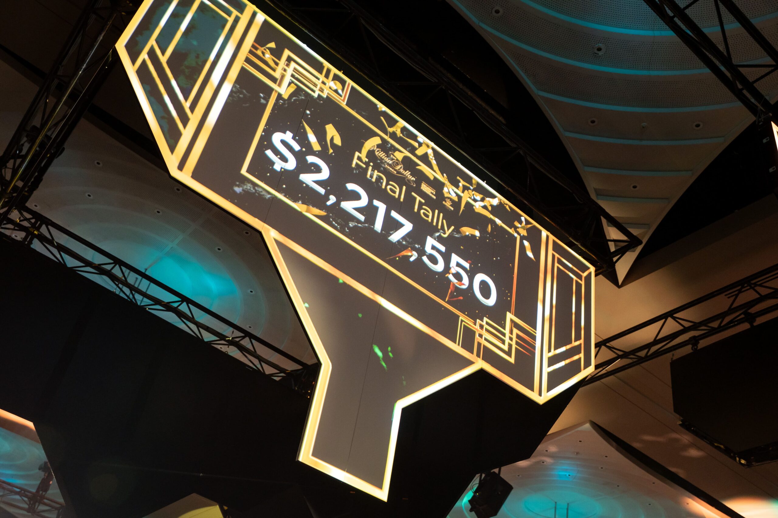 A large illuminated sign displays Final Tally: $2,217,550 in bold white text, hanging from the ceiling at the Million Dollar Lunch 2025 event. The sign features a geometric, art deco-inspired design.