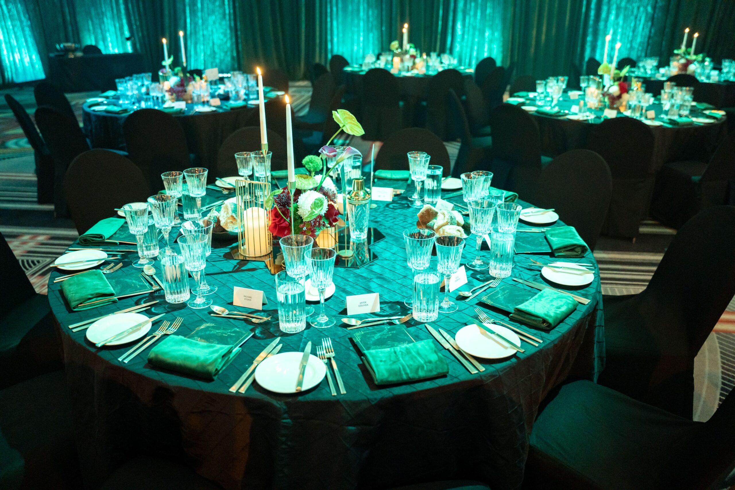 A round banquet table set for a formal event at Million Dollar Lunch features green napkins, glassware, plates, silverware, candles, and a floral centerpiece. Other elegantly arranged tables glow under teal lighting in the background.
