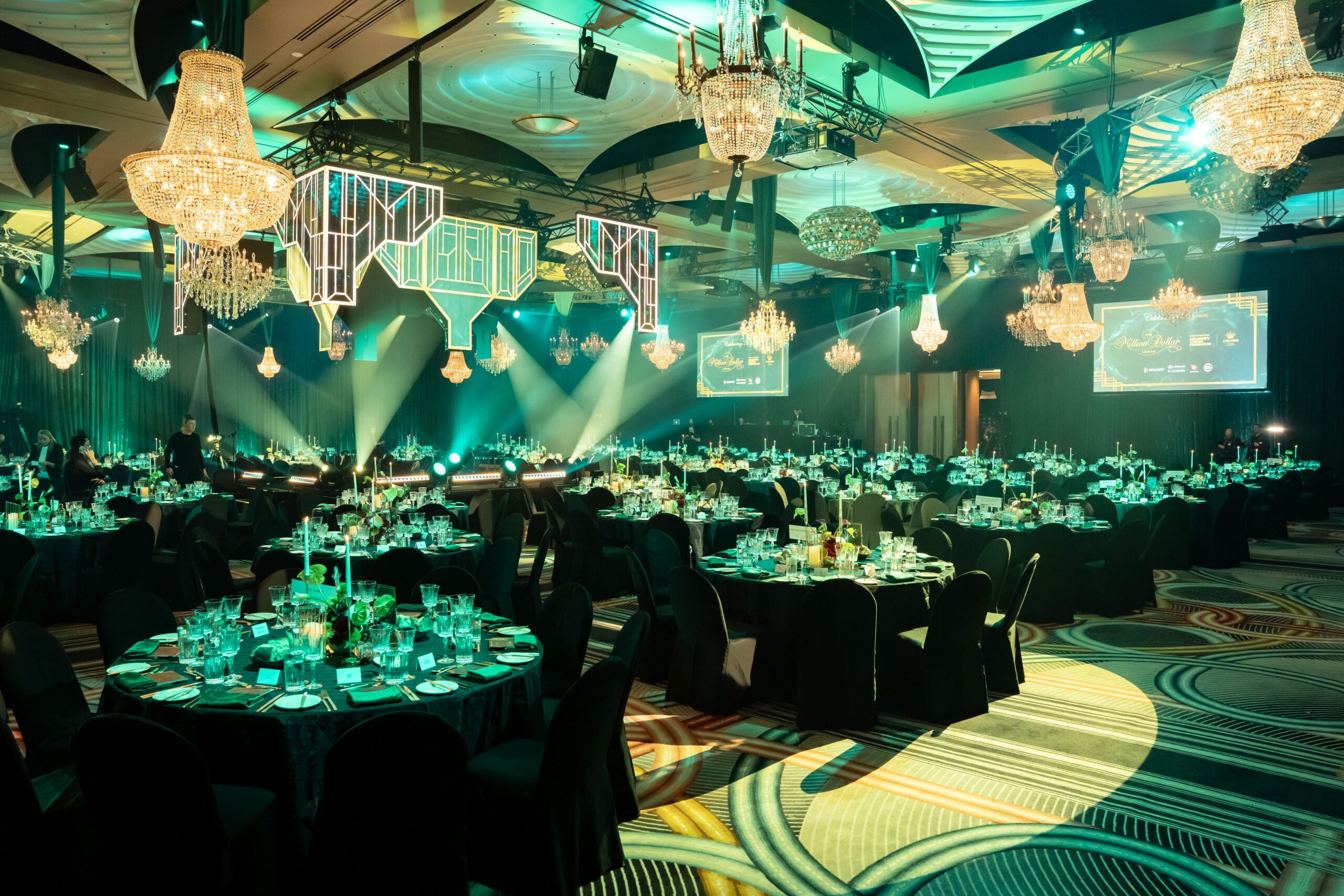 A large, elegant ballroom is set for Million Dollar Lunch, with round tables covered in black cloths, chandeliers, dramatic green lighting, and Art Deco decorations hanging from the ceiling.