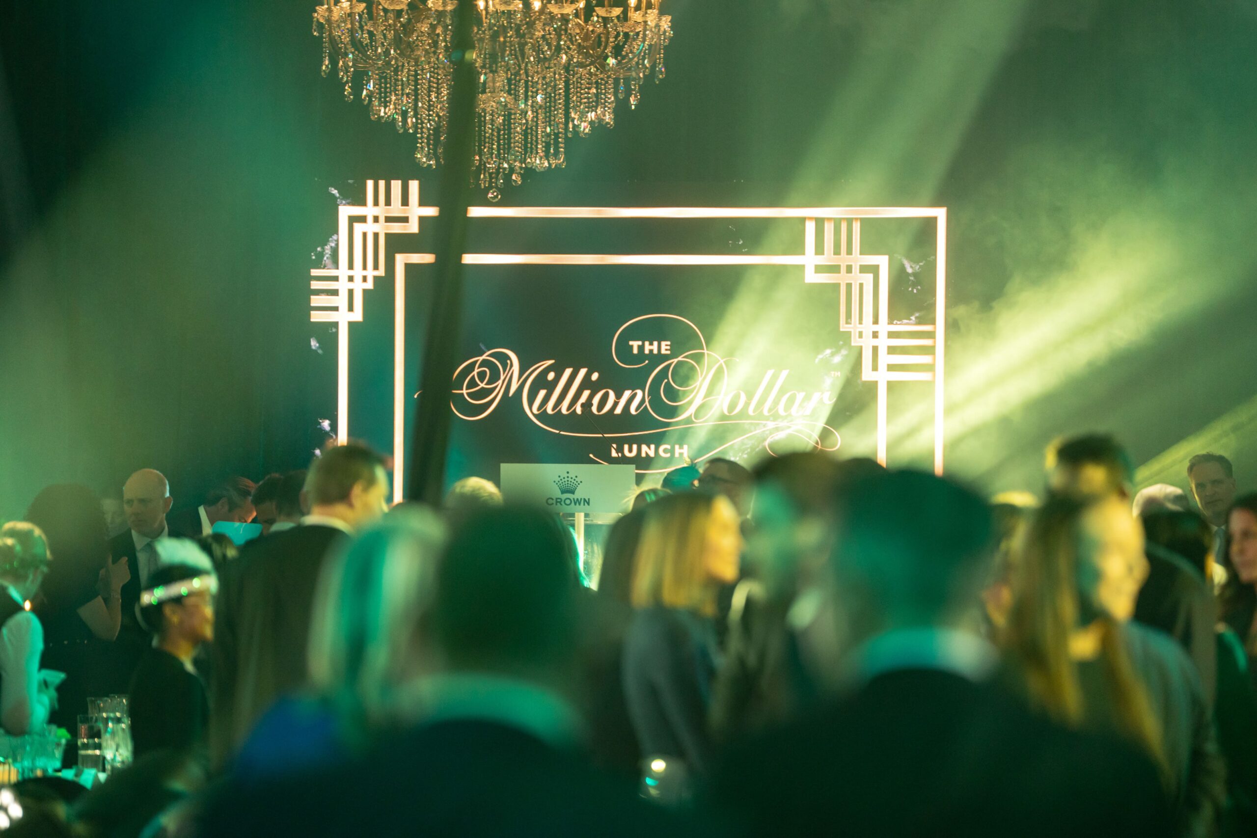 A crowd of people in formal attire gathers at an elegant Million Dollar Lunch event under a chandelier, with a brightly lit sign in the background reading “The Million Dollar Lunch.”.