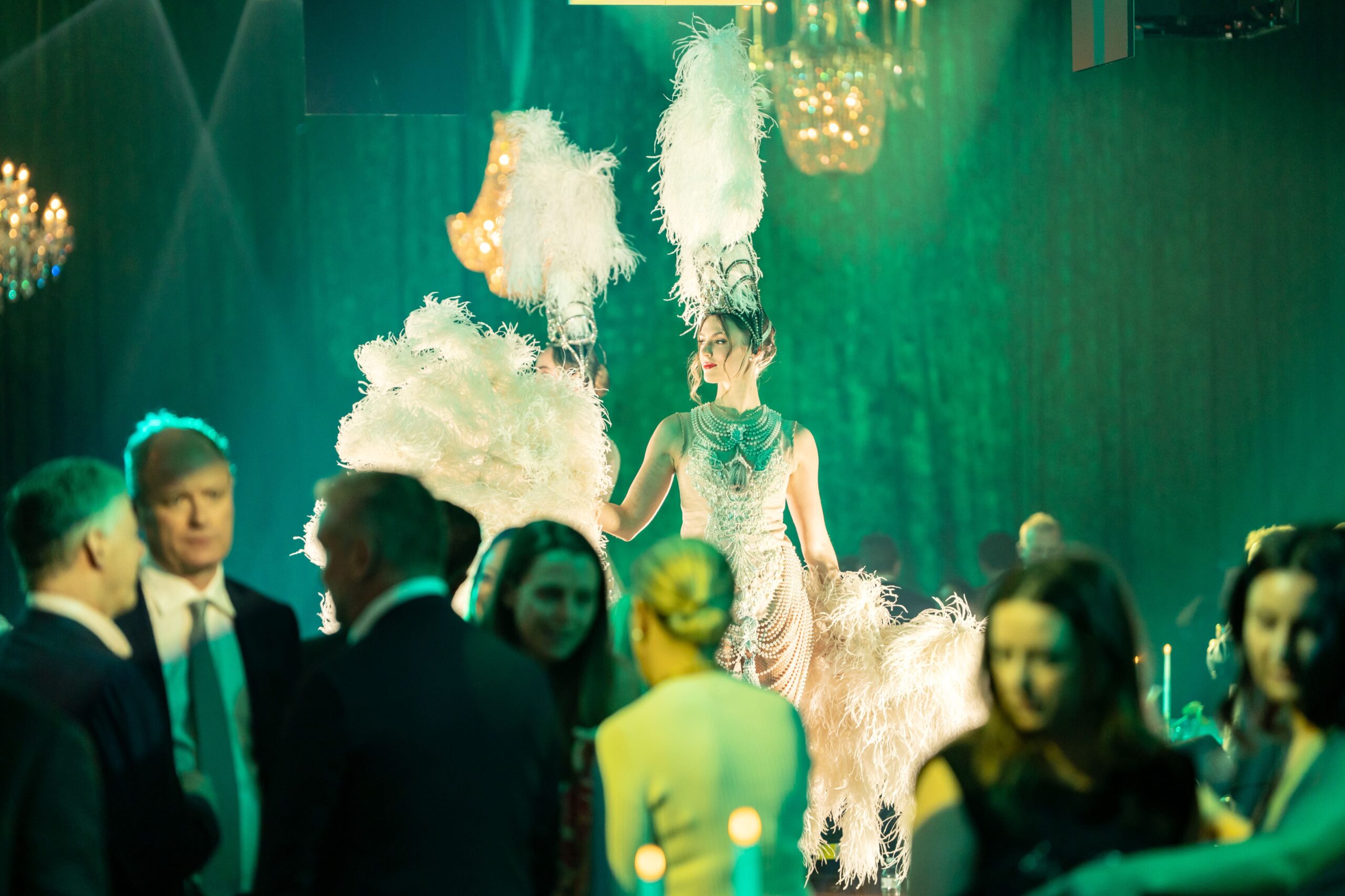 A performer in a glamorous, feathered costume stands under green lighting on stage at Million Dollar Lunch, surrounded by people in formal attire at an elegant event. Chandeliers hang from the ceiling in the background.