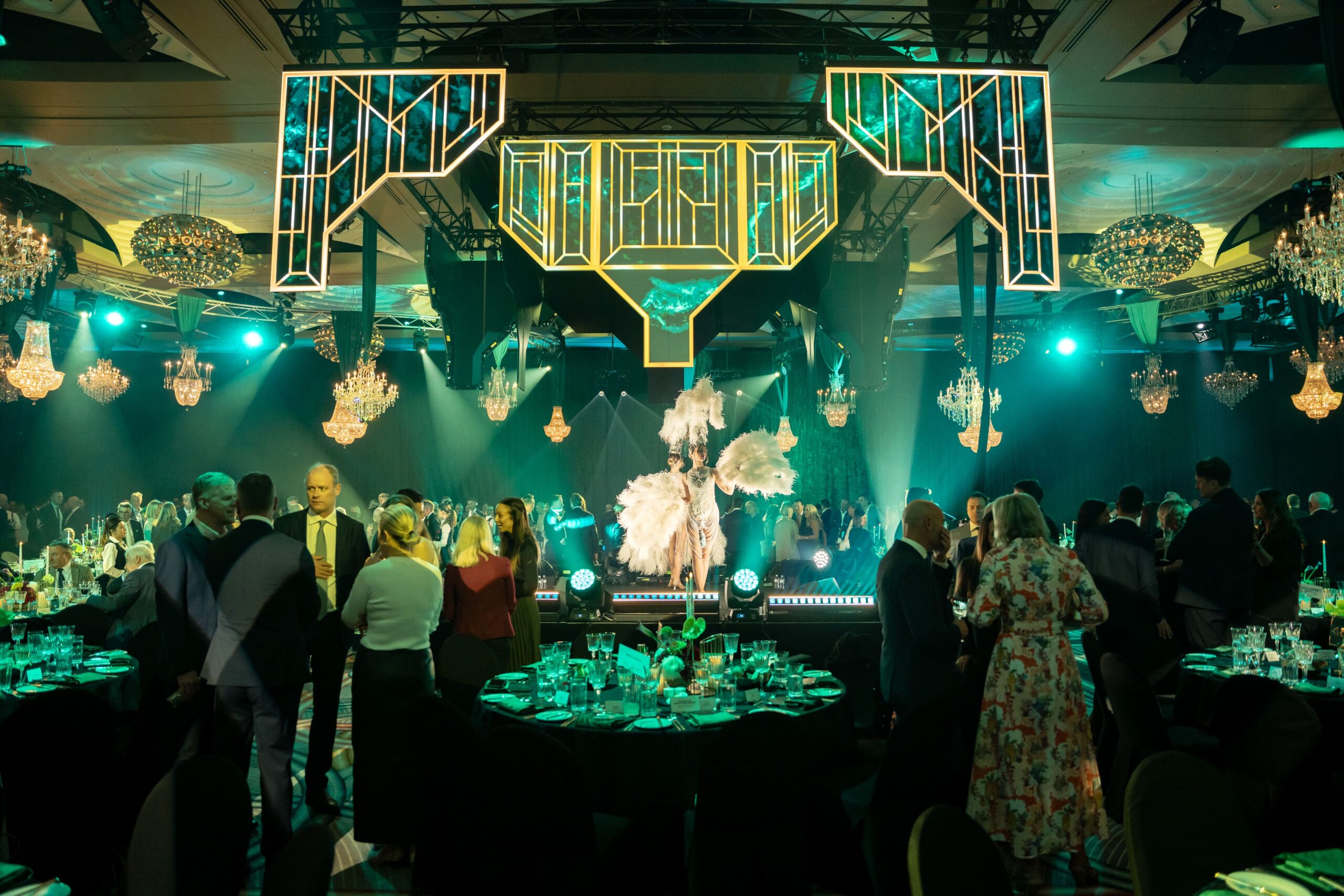A glamorous Million Dollar Lunch event with guests in formal attire, round dinner tables, and chandeliers; a performer in a feathered costume entertains on a stage with art deco-style lighting in green and gold.