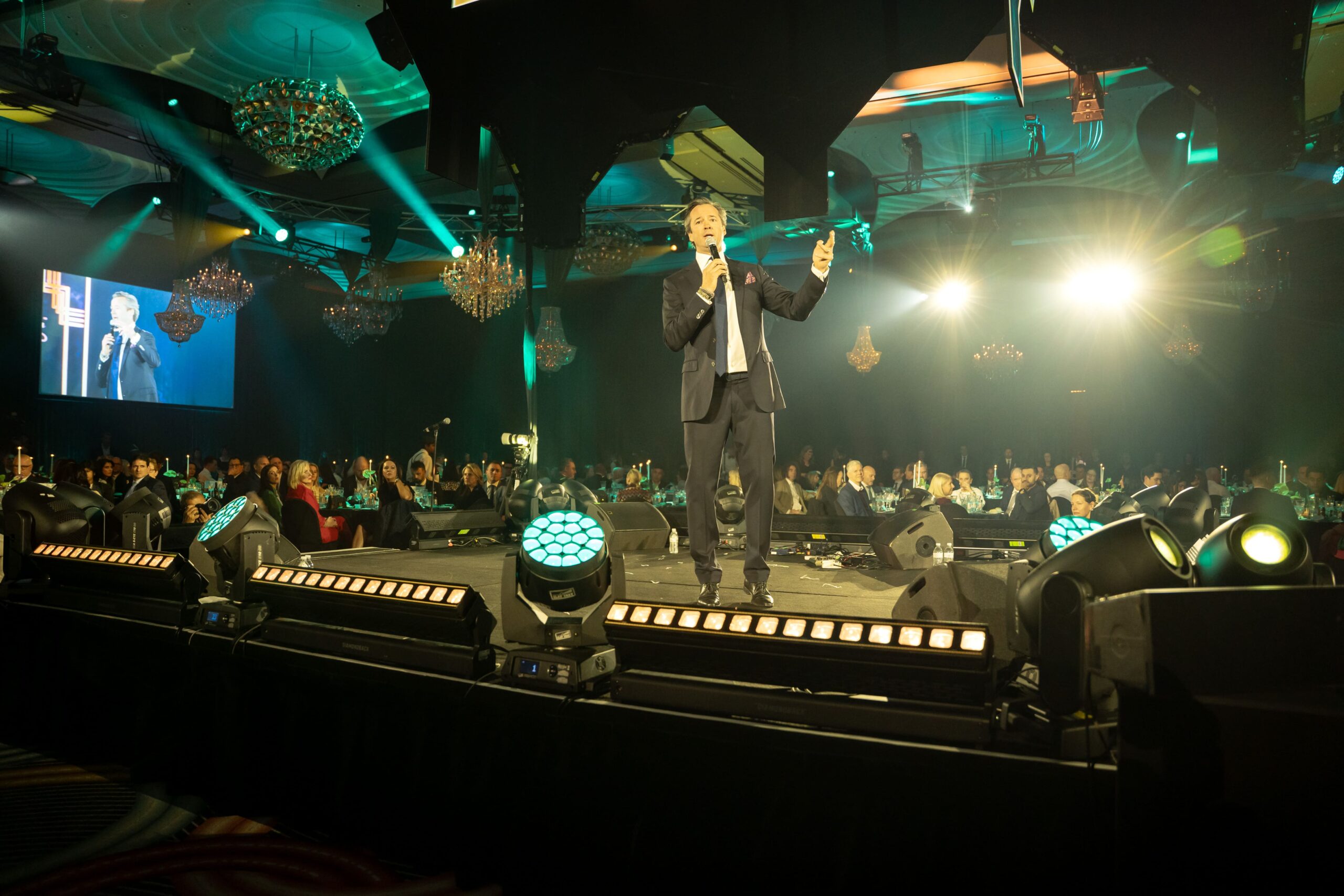 A man in a suit speaks on stage with a microphone at a large indoor event. Bright stage lights shine toward the audience, and people are seated at tables. An event screen behind him displays his image at Million Dollar Lunch 2025.