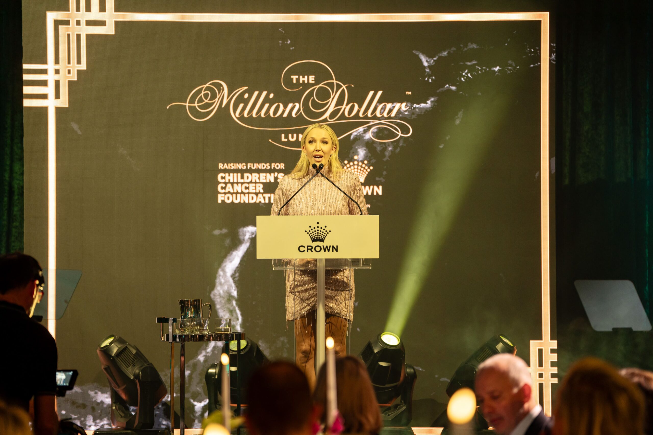 A woman stands at a podium labeled Crown, speaking at the Million Dollar Lunch, an Million Dollar Lunch 2025 event raising funds for the Children’s Cancer Foundation. Elegant decor and lights highlight the stage, with audience members visible in the foreground.