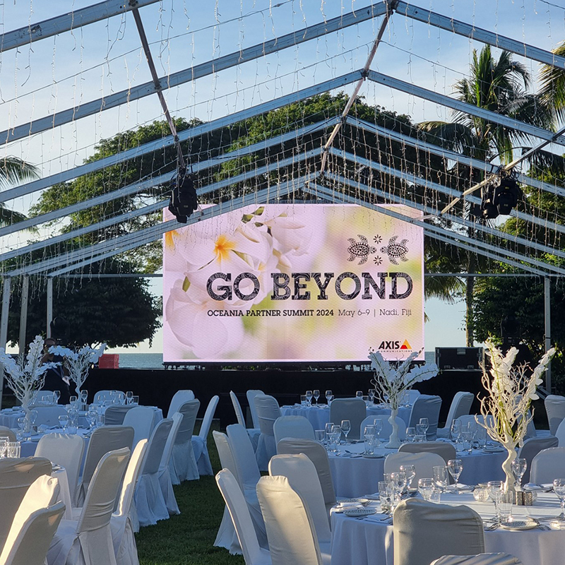 A large outdoor event tent with tables and chairs set for a formal gathering, produced by a leading event production company in Fiji. A big screen displays GO BEYOND, Oceania Partner Summit 2024. Palm trees and blue sky complete the setting.
