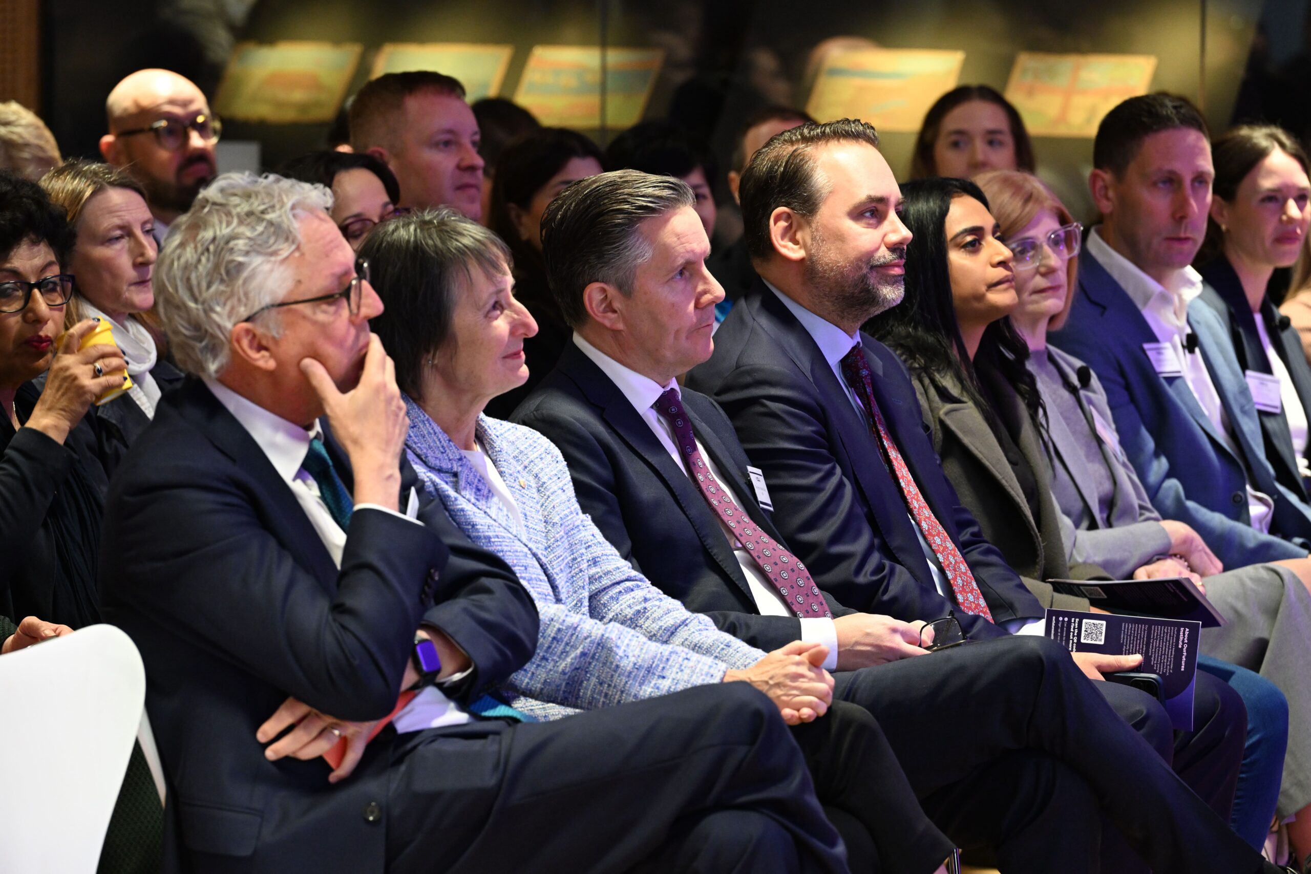 비즈니스 복장을 한 사람들이 ADIA 2023에서 일렬로 나란히 앉아 행사나 프레젠테이션을 주의 깊게 지켜보고 있습니다. 몇몇은 서류를 들고 있으며 표정은 진지하고 몰입되어 있습니다.