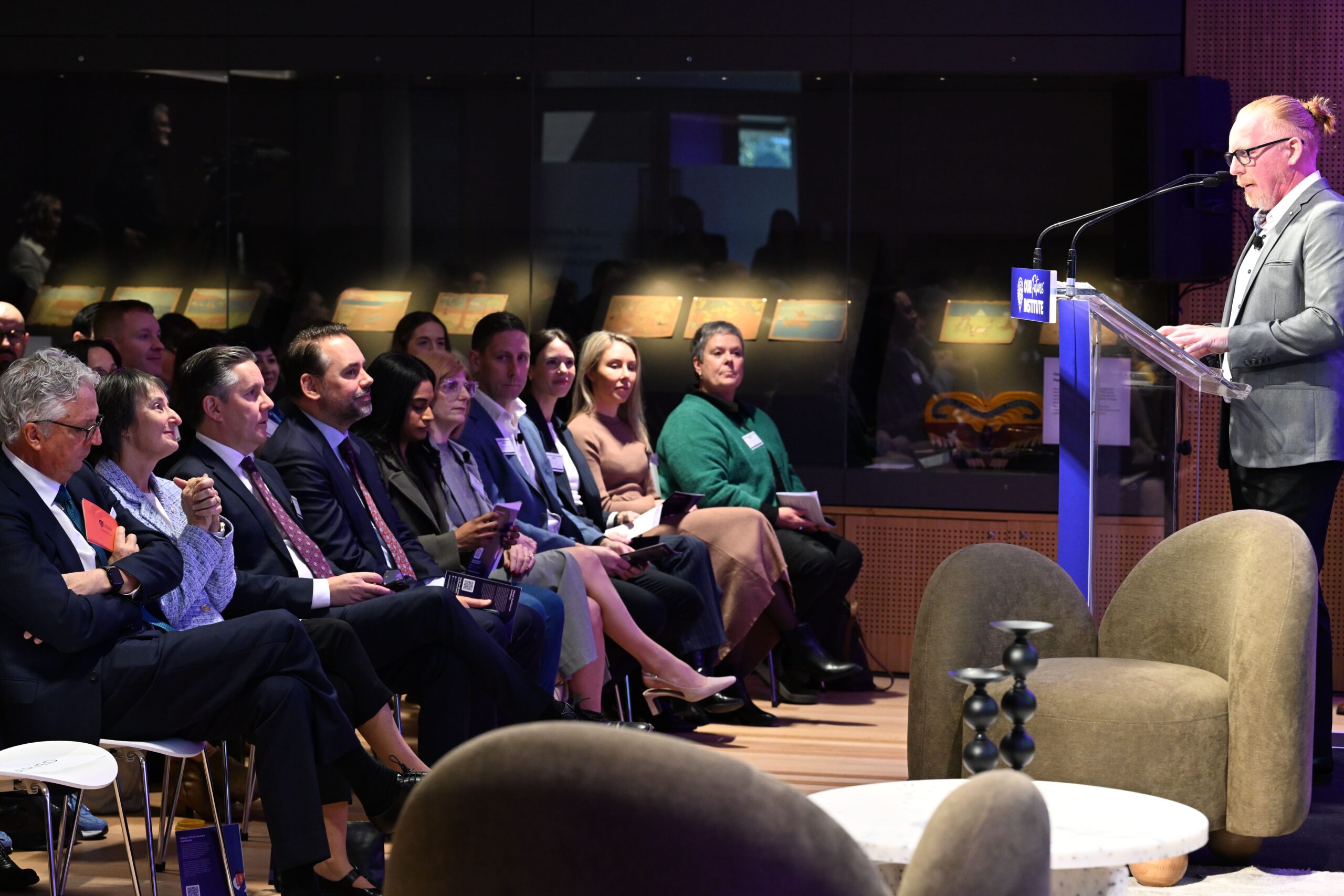 조명이 밝은 현대식 회의실에서 한 연사가 ADIA 2023에 모인 청중에게 연설하고 있습니다. 비즈니스 복장을 한 참석자들은 조명이 켜진 진열장이 배경에 보이는 가운데 귀를 기울이고 있습니다.