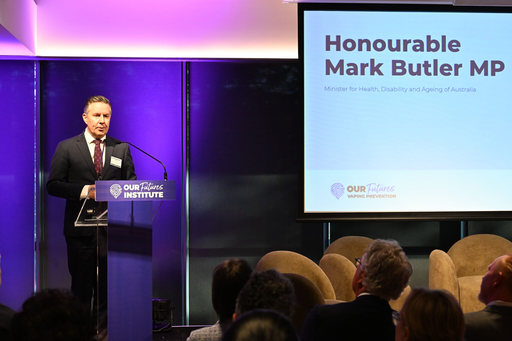 정장을 입은 한 남성이 ADIA 2023의 공식 행사 중 연단에서 연설하고 있습니다. 그 옆의 스크린에 텍스트가 표시됩니다: 명예로운 마크 버틀러 의원, 호주 보건, 장애 및 고령화부 장관, 우리 미래 연구소.