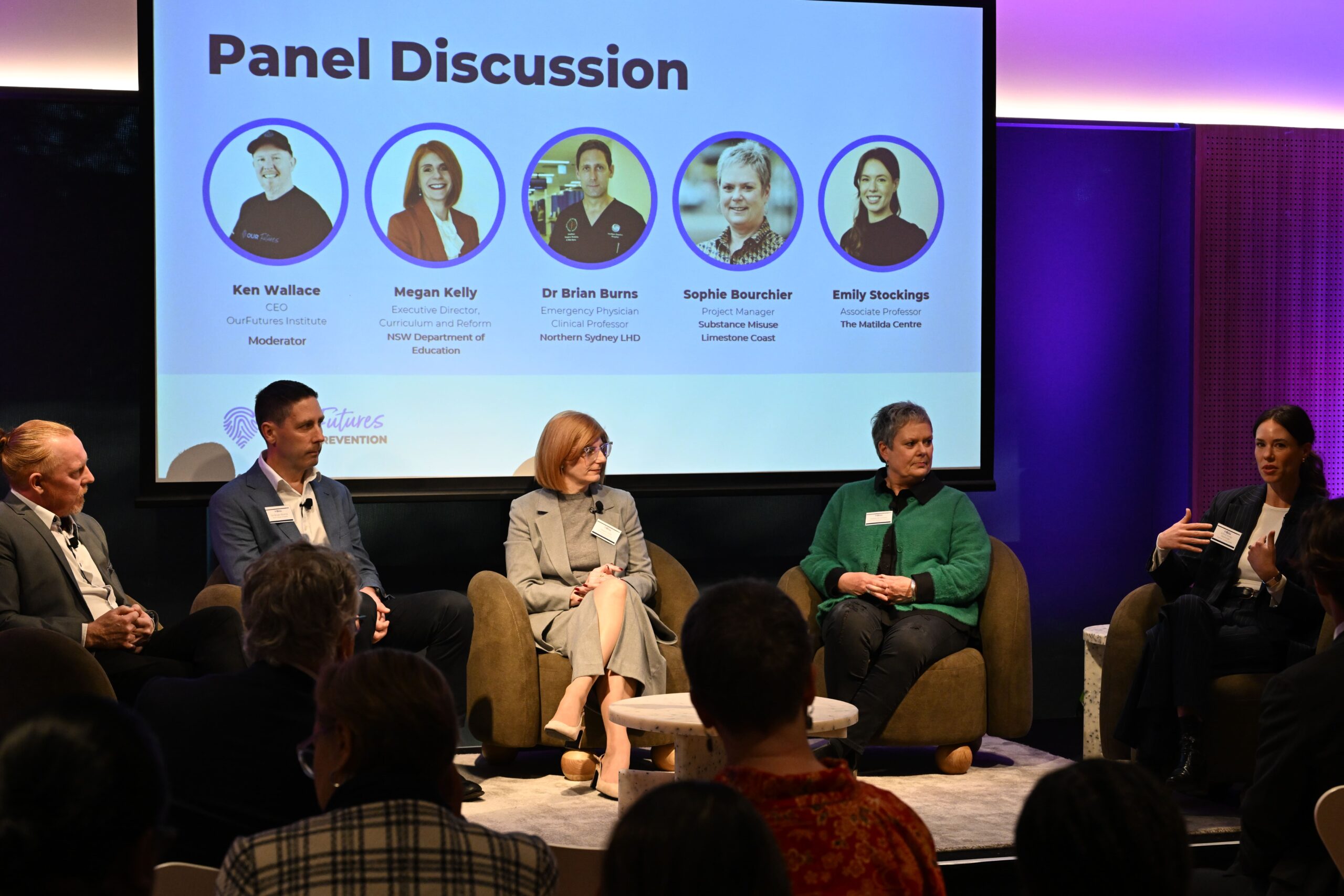 ADIA 2023 토론이 진행되는 동안 5명의 패널이 무대에 앉아 있고, 그 뒤에 스크린에 패널의 이름과 역할, 사진이 표시됩니다. 청중은 한 패널이 손으로 제스처를 취하며 말하는 것을 주의 깊게 경청합니다.