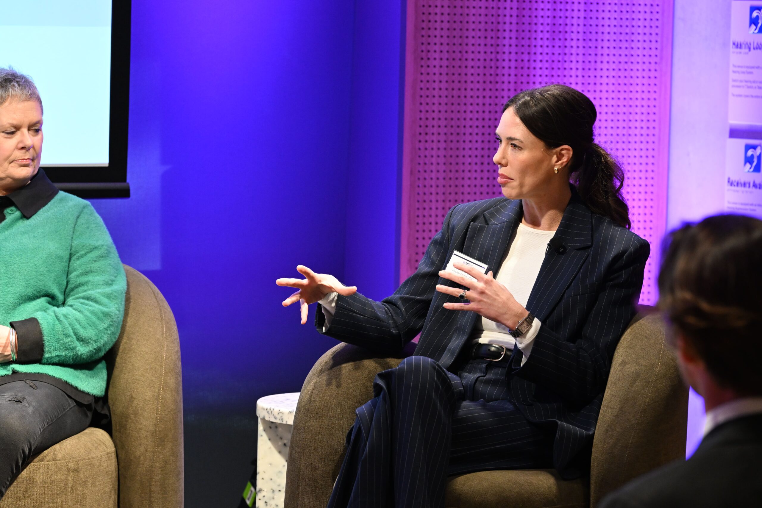 어두운 핀스트라이프 정장을 입은 한 여성이 ADIA 2023 패널 토론에서 발언하는 동안 제스처를 취하고 있습니다. 그녀는 보라색 조명이 켜진 배경과 프로젝션 스크린이 부분적으로 보이는 갈색 의자에 앉아 다른 두 사람을 마주보고 있습니다.