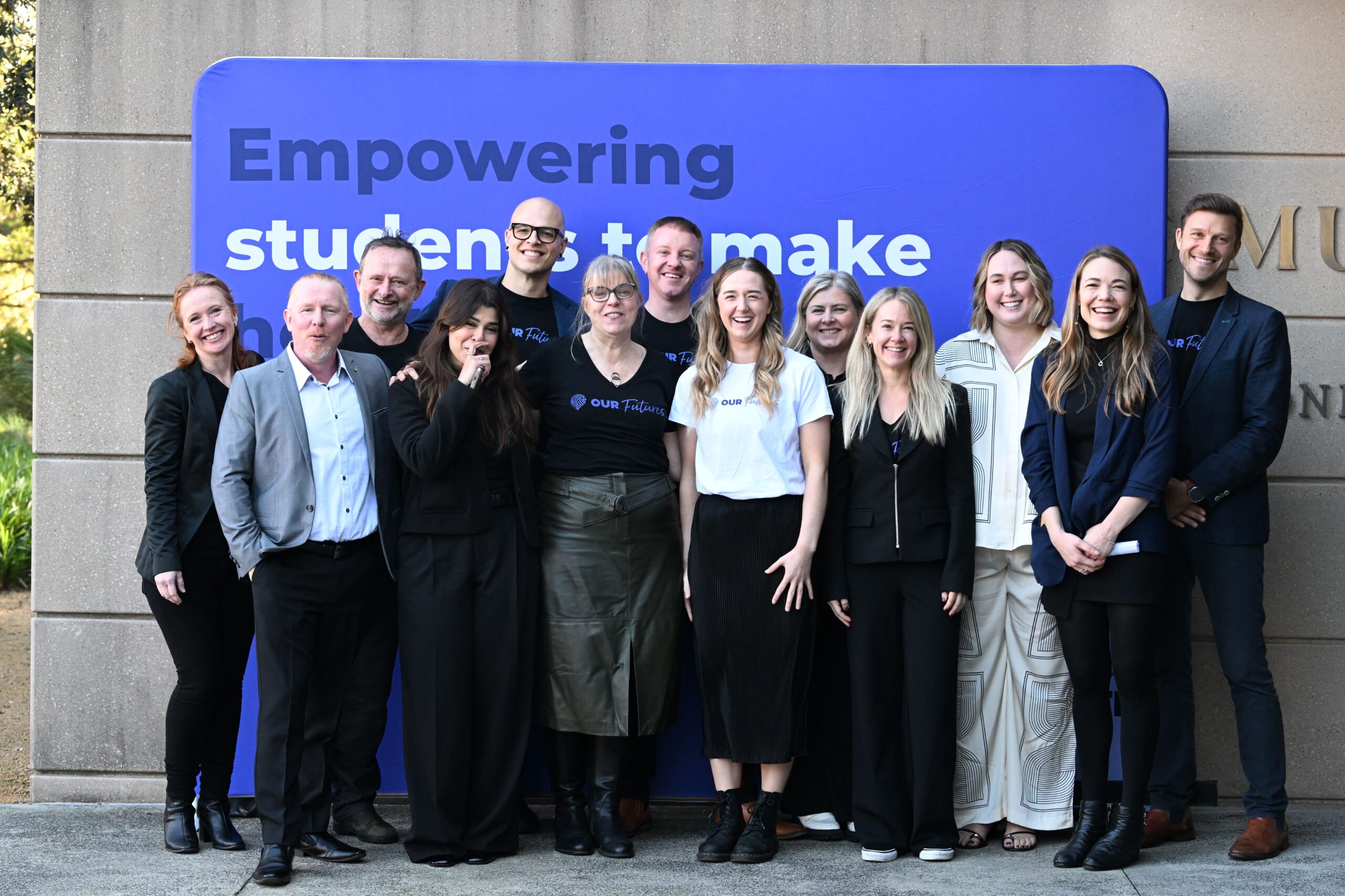 13명의 사람들이 '학생들의 제작 역량 강화'라고 적힌 커다란 파란색 간판 앞에서 웃으며 서 있습니다. 비즈니스 캐주얼 복장을 한 이들은 행복하고 단합된 모습으로 ADIA 2023에서 거둔 성과를 축하하고 있습니다.