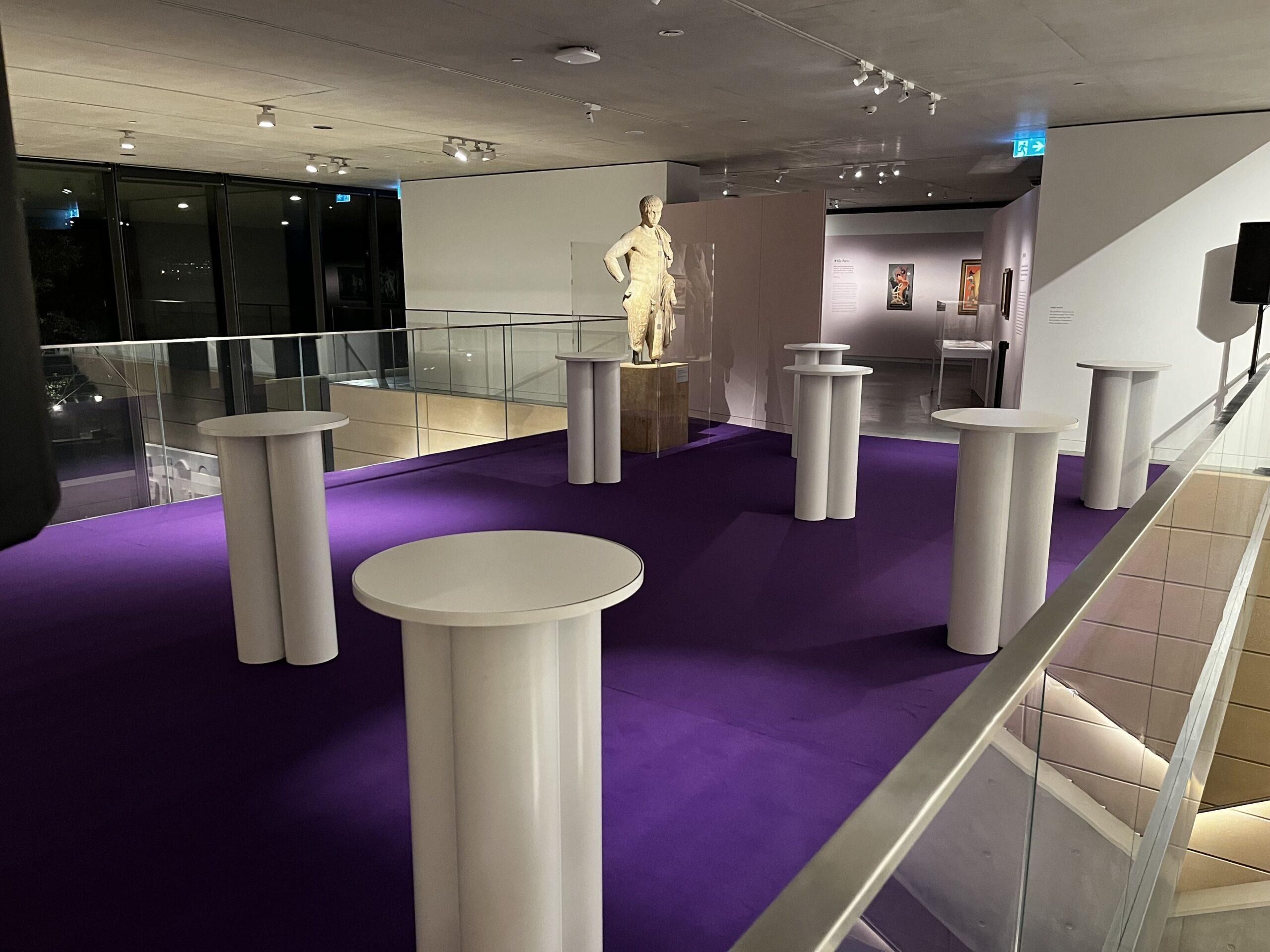 보라색 카펫이 깔린 현대적인 갤러리 공간으로, 고전적인 입상 조각상을 중심으로 여러 개의 키가 큰 흰색 원형 테이블이 배치되어 있습니다. 조명이 밝고 예술 작품이 전시되어 있는 이 공간은 ADIA 2023 행사를 위해 마련된 공간입니다.