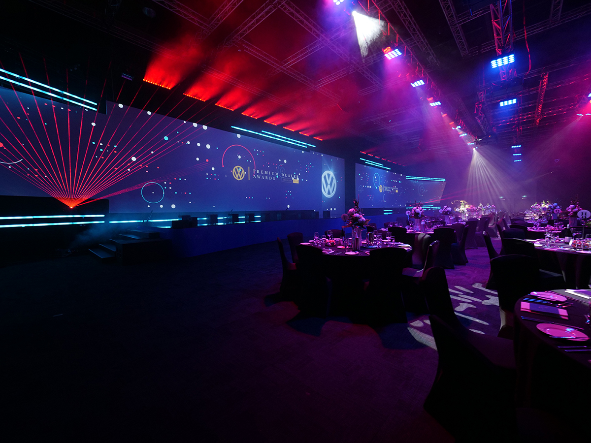 A large, dimly lit event hall hosting a conference and gala dinner, with round tables set for dinner, dramatic stage lighting, and colorful digital graphics on wide screens featuring the Volkswagen logo and futuristic designs.
