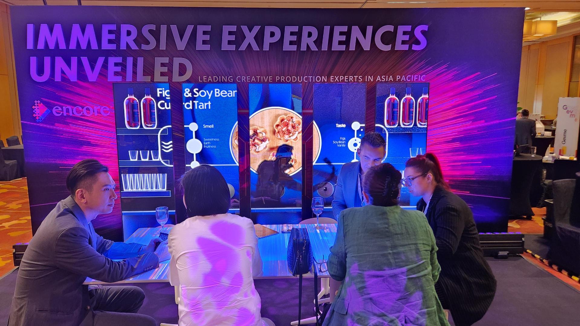 Four people sit at a table discussing in front of a colorful display that reads Immersive Experiences Unveiled, with lighting effects and creative production visuals in a conference or event setting.