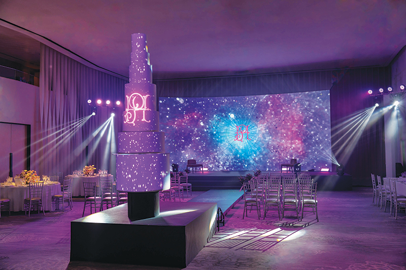 A large, modern indoor event space perfect for weddings, with a tall illuminated cake, round tables with chairs, and a stage. Purple and blue lights create a starry effect on the walls, adding to the festive, elegant atmosphere.