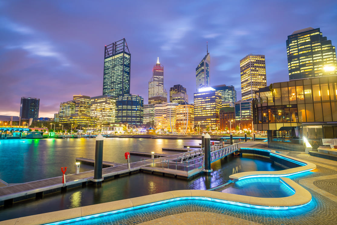 A vibrant city skyline at dusk with illuminated skyscrapers reflecting on the water, modern waterfront walkways, and glowing blue lights outlining the paths in the foreground.