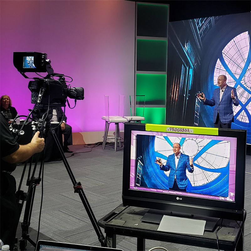 A man in a blue suit speaks on stage in front of a large screen with a circular design, while a camera films him and his image appears on a monitor in the foreground. Other people sit nearby.