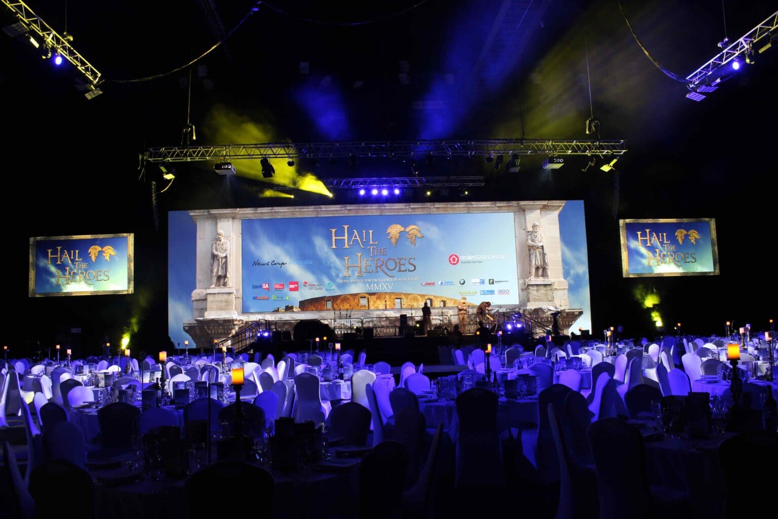 A grand ballroom set for an event, with round tables covered in white cloths and chairs facing a brightly lit stage displaying “HALL OF HEROES” on a large screen, with two smaller screens showing the same text.