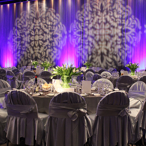 A formal event space suitable for various event types, featuring round tables with gray tablecloths and matching chair covers, elegant glassware and flowers, purple uplighting, and decorative wall projections in the background.
