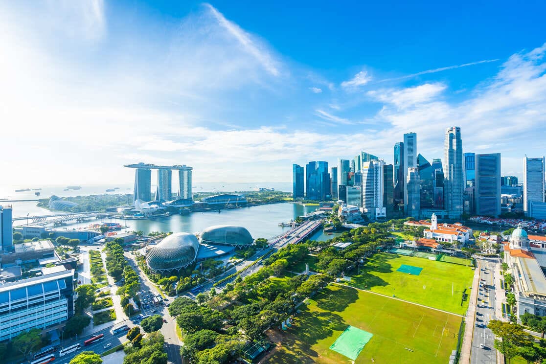 Aerial view of Singapore’s Marina Bay area, featuring modern skyscrapers, the Marina Bay Sands hotel, waterfront, green parks, and sports fields under a bright blue sky.