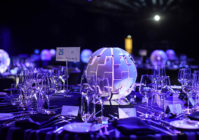 A decorated banquet table set for an event, featuring a glowing, spherical centerpiece with geometric patterns, surrounded by glassware and place cards, all illuminated by violet and blue lighting.