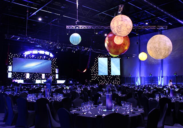 A large event hall with round tables set for dinner, blue lighting, and large hanging planets as decorations. Two big screens and stage lights are visible in the background.