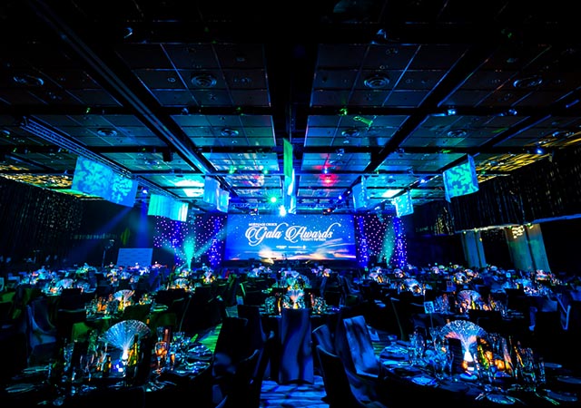 A large, elegant banquet hall set for a gala event, with round tables, glowing centerpieces, and blue ambient lighting. A stage at the front has “Gala Awards” displayed on a screen.