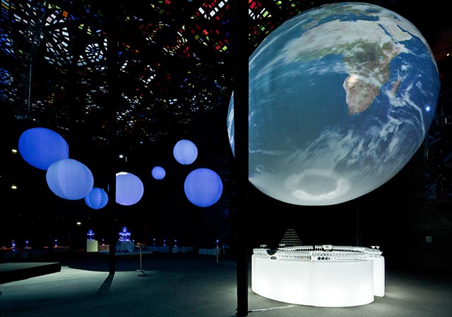 A dark exhibition space displays large illuminated globes, with one showing Earths surface in detail and others glowing in blue hues, beneath a stained glass ceiling. White display tables with small objects are arranged below.