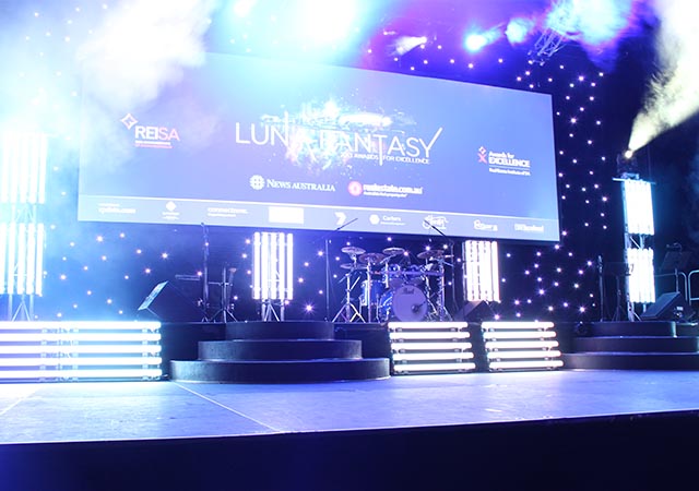 A brightly lit stage with spotlights, LED lights, and a large screen displaying “Luna Fantasy” and event sponsors, set against a backdrop of small star-like lights. Drums and other instruments are positioned on stage.