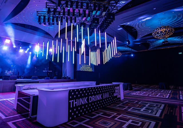 A modern event venue with a large bar labeled “THINK DRINK BAR,” colorful hanging light tubes, and a stage with spotlights. The room is bathed in blue and purple lighting, with patterned carpeting and decorative ceilings.