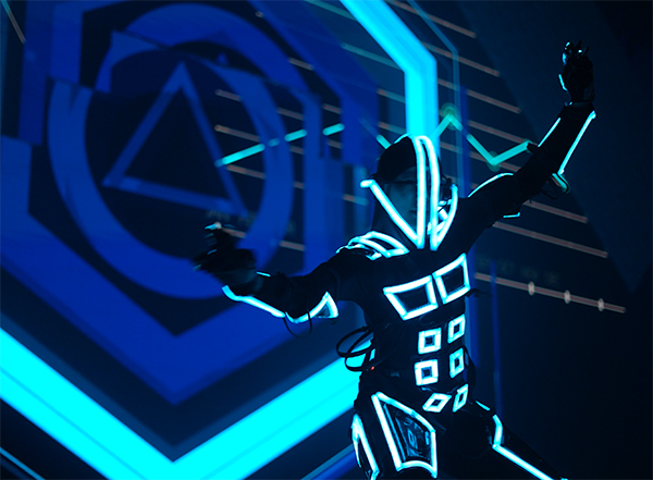 A person in a futuristic suit with glowing neon blue lines poses energetically in front of abstract blue geometric shapes and patterns.