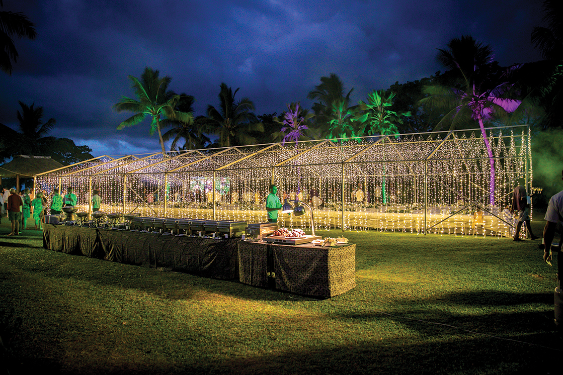 An outdoor evening event with a large illuminated canopy of string lights, palm trees in the background, and buffet tables on a grassy lawn—expertly designed by an event production company in Fiji, as people gather under the glowing lights.