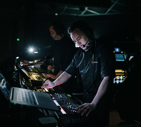 Two people wearing headsets operate sound and lighting equipment in a dark control room, illuminated by screens and small lights, suggesting they are managing audio and visuals for a live event or production.