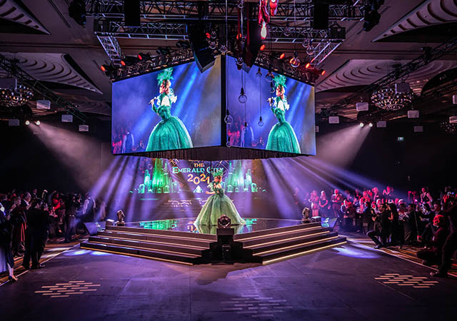 A performer in a green dress stands on a stage under dramatic lighting, with large screens above—showcasing event technology—displaying her image. The audience surrounds the stage in a spacious, decorated event hall.