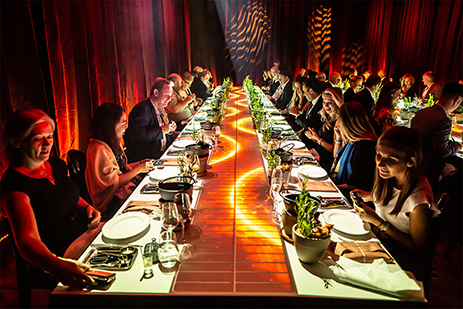 A long, glowing dinner table with people seated on both sides, enjoying a lively encore meal in a dimly lit room with red and orange lighting effects on the walls. The scene is both elegant and vibrant.