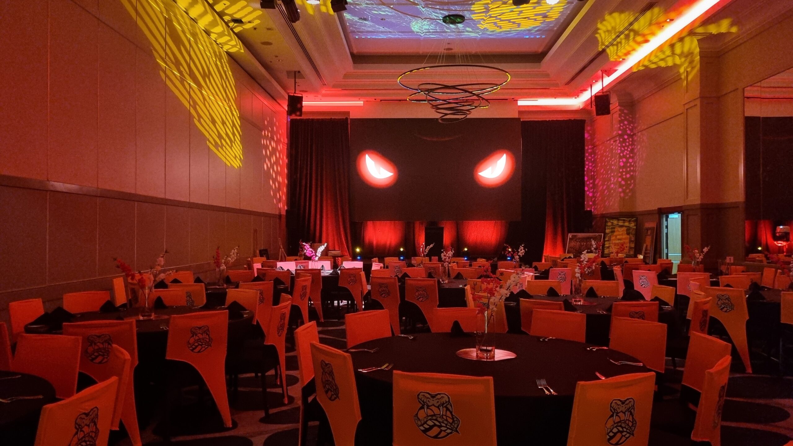 A large event room with round tables and orange chairs, decorated with fierce stylized faces for the Taipans' Gala Dinner 2025. The stage features two glowing red eyes, with red and yellow lights creating dramatic patterns on walls and ceiling.