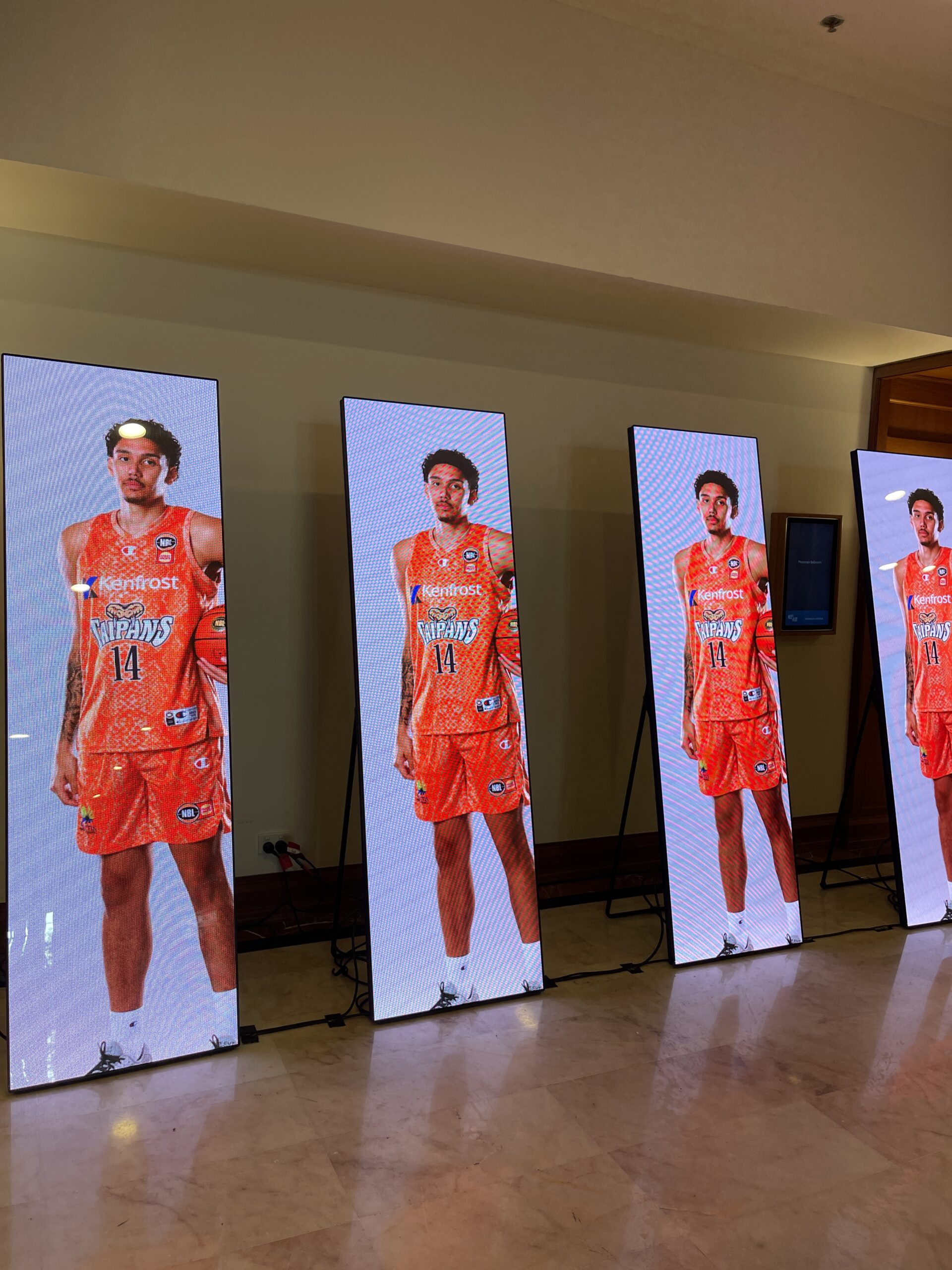 Four tall, vertical digital screens display a basketball player in an orange uniform with the number 14, posing against a white background. The setup, on a marble floor indoors, showcases highlights for the Taipans' Gala Dinner 2025.