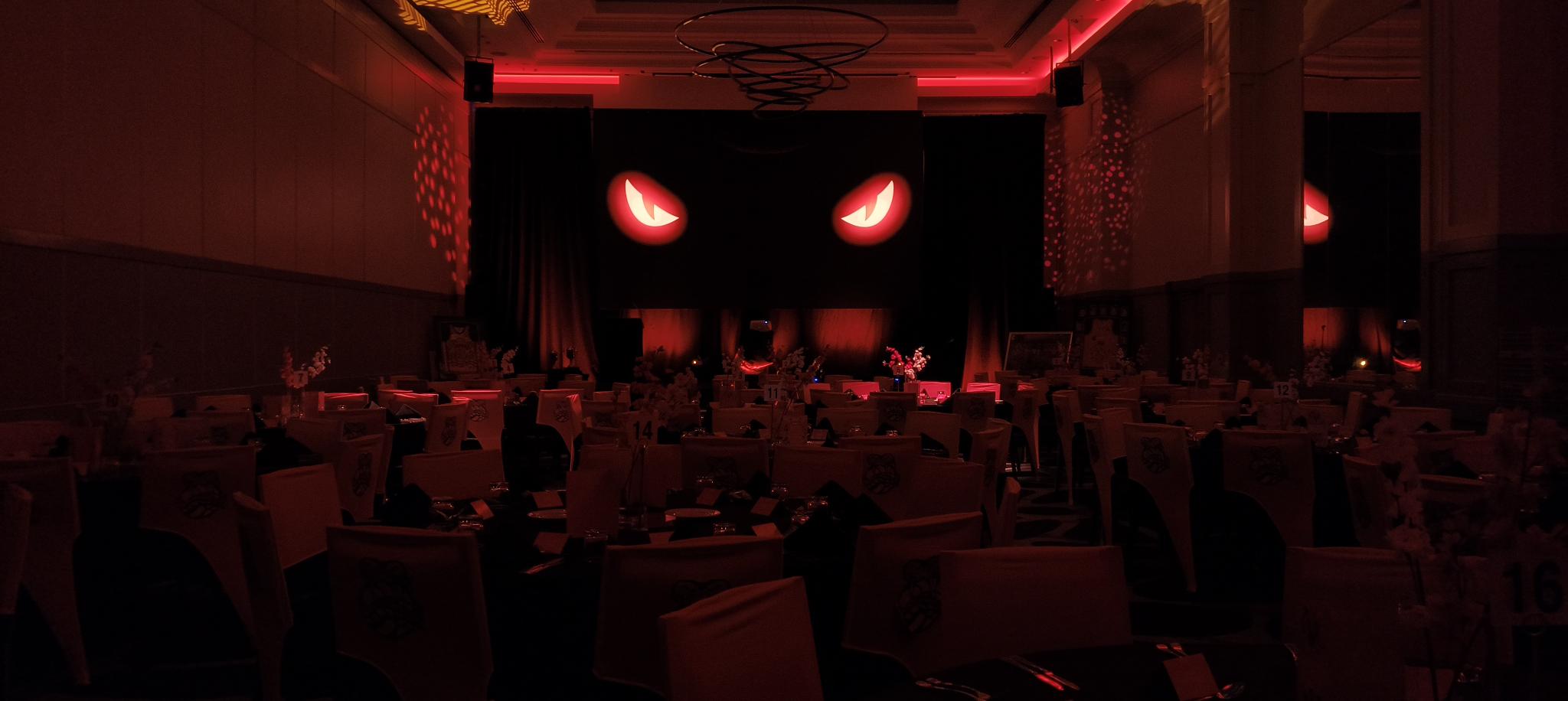 A dimly lit event hall with tables and chairs, illuminated by red lighting. The main feature is a stage with a backdrop displaying large, glowing red eyes, setting a dramatic and mysterious mood for the Taipans' Gala Dinner 2025.