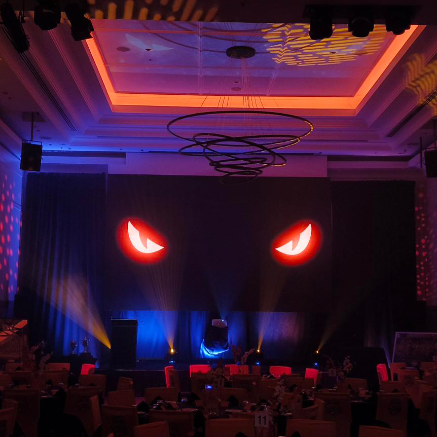 A dimly lit event hall by Encore Cairns features a large screen displaying two glowing, red, cartoonish eyes. Colorful stage lights and spiral ceiling decor add to the dramatic, mysterious atmosphere crafted by this top event company Cairns.