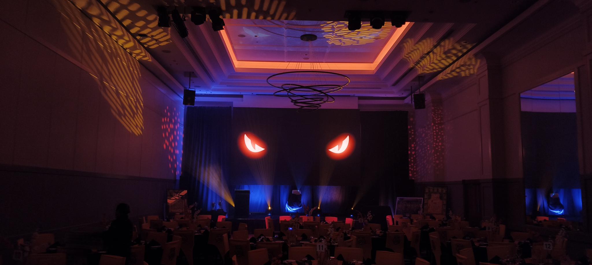 A dark event hall with dramatic red and yellow lighting hosts the Taipans' Gala Dinner 2025. Large, glowing, menacing eyes loom on the stage backdrop, while round tables with chairs await guests for an unforgettable night.