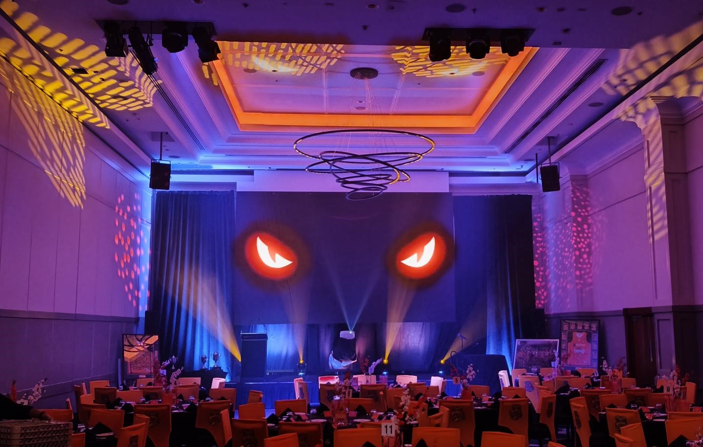 A decorated banquet hall with orange and purple lighting sets the scene for the Taipans' Gala Dinner 2025. Tables draped in orange cloths and a stage with a large screen of glowing, sinister eyes add drama beneath dynamic spotlights.