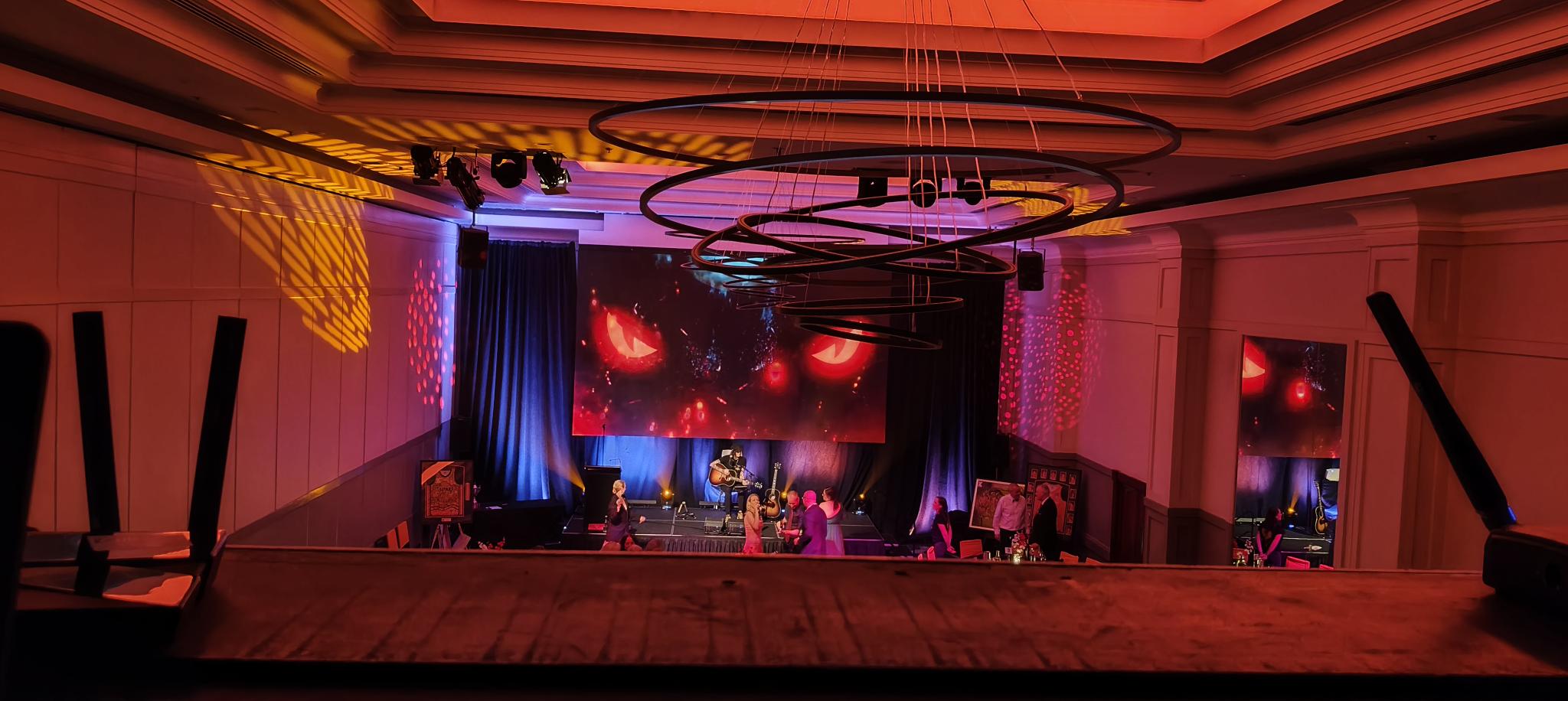 A dimly lit event room with red lighting sets the scene at Taipans' Gala Dinner 2025, where two people are seated on stage in conversation. A large screen behind them displays red, glowing eyes, and circular light fixtures hang from the ceiling.