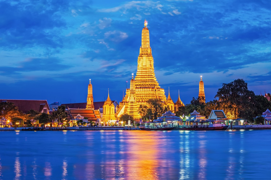 Wat Arun temple in Bangkok, Thailand, illuminated at dusk with golden lights reflecting on the Chao Phraya River, set against a vibrant blue sky with scattered clouds.