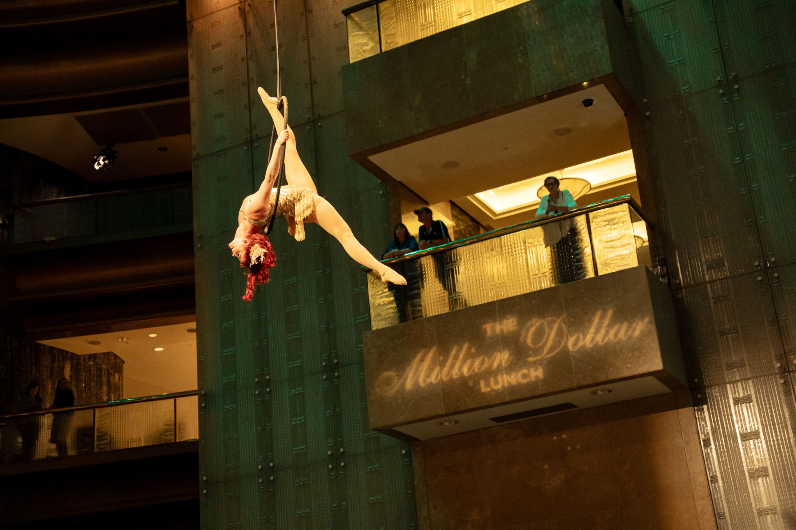 Aerial performer with red hair and a sparkling outfit hangs upside down from a rope in a theater during Million Dollar Lunch 2025, while people watch from a balcony above a sign that reads The Million Dollar Lunch.