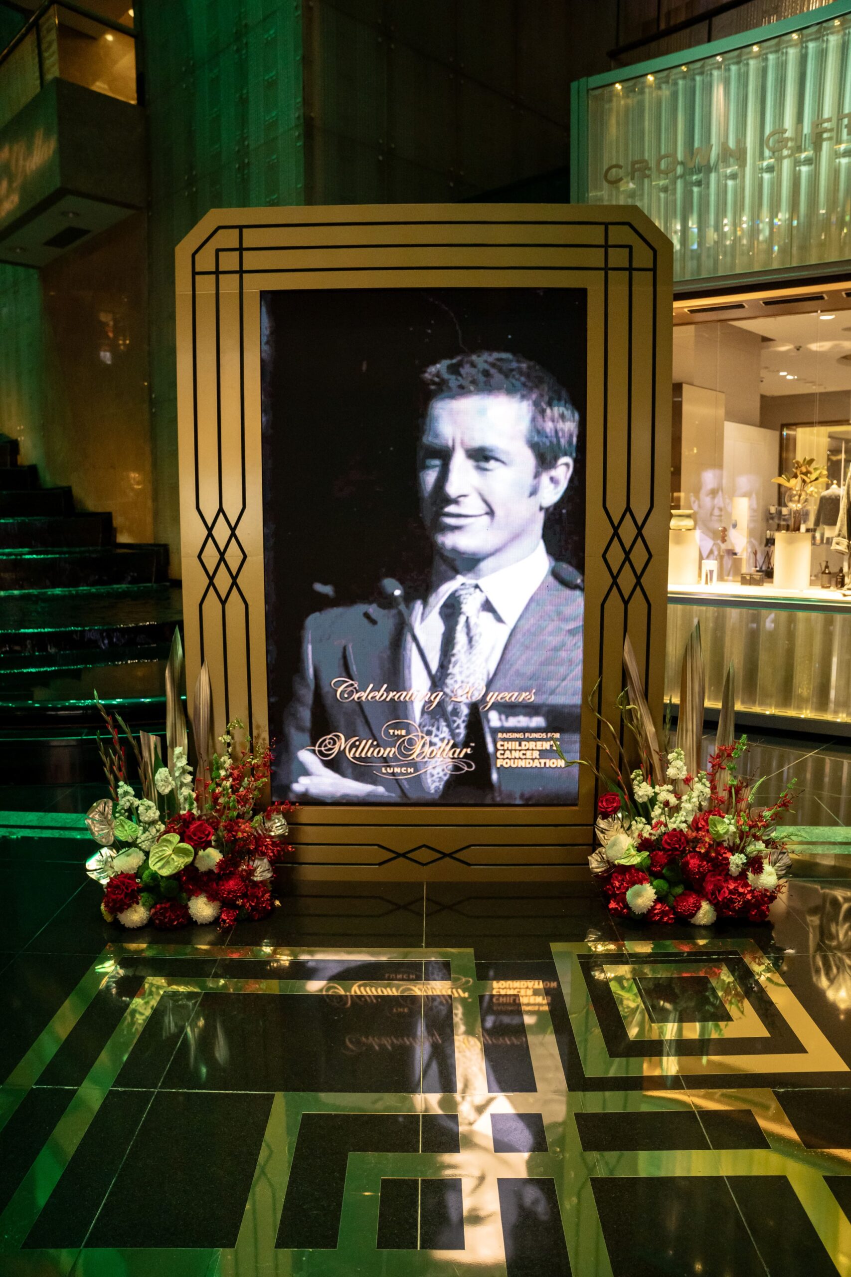 A large digital screen at Million Dollar Lunch displays a black-and-white portrait of a smiling man in a suit, framed by gold, Art Deco-style borders. Red and white flower arrangements sit at the screen’s base on a shiny black floor.