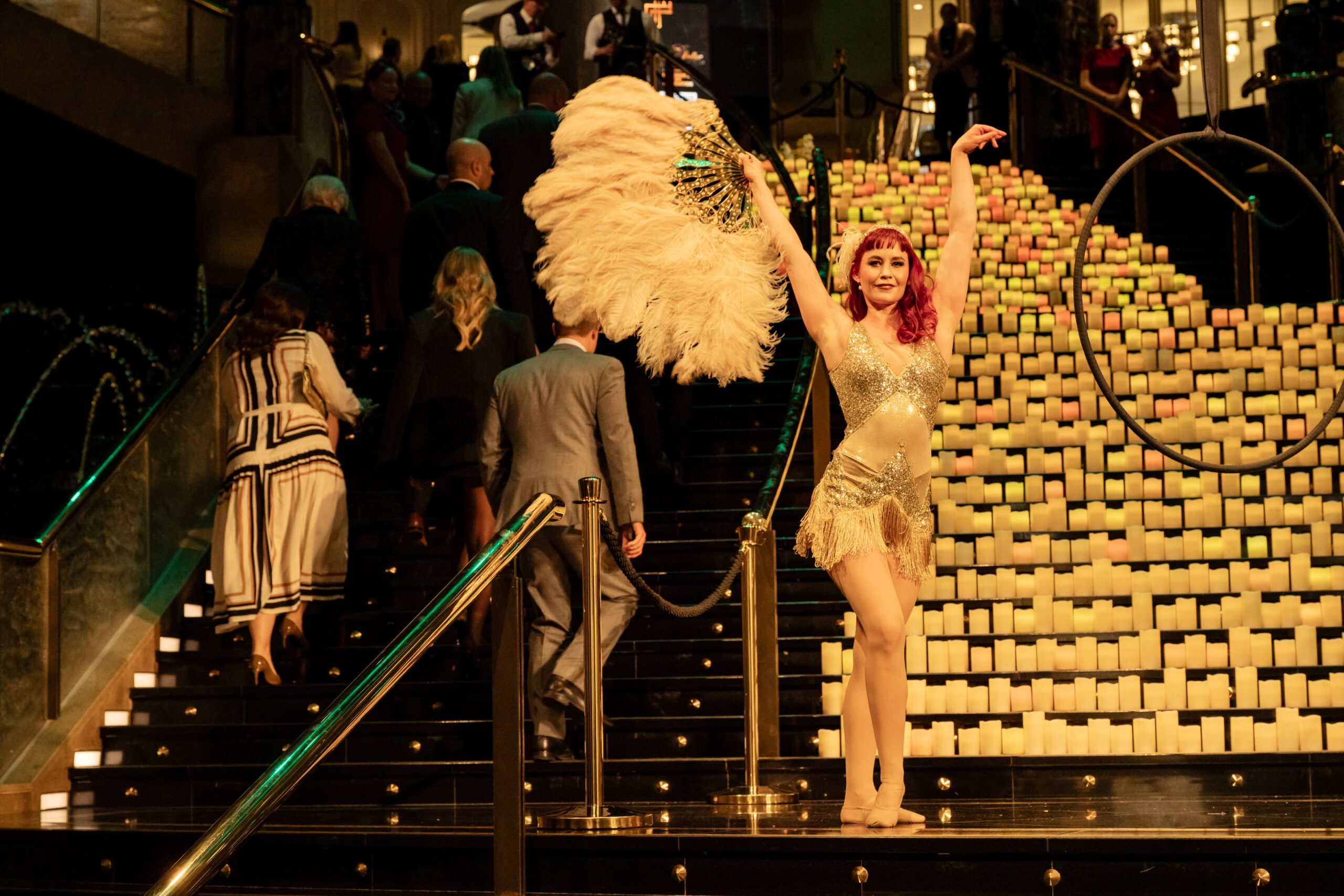 A performer in a sparkling costume and feathered headpiece poses on a staircase lined with glowing candles at Million Dollar Lunch 2025, while people in formal attire ascend the stairs behind her.