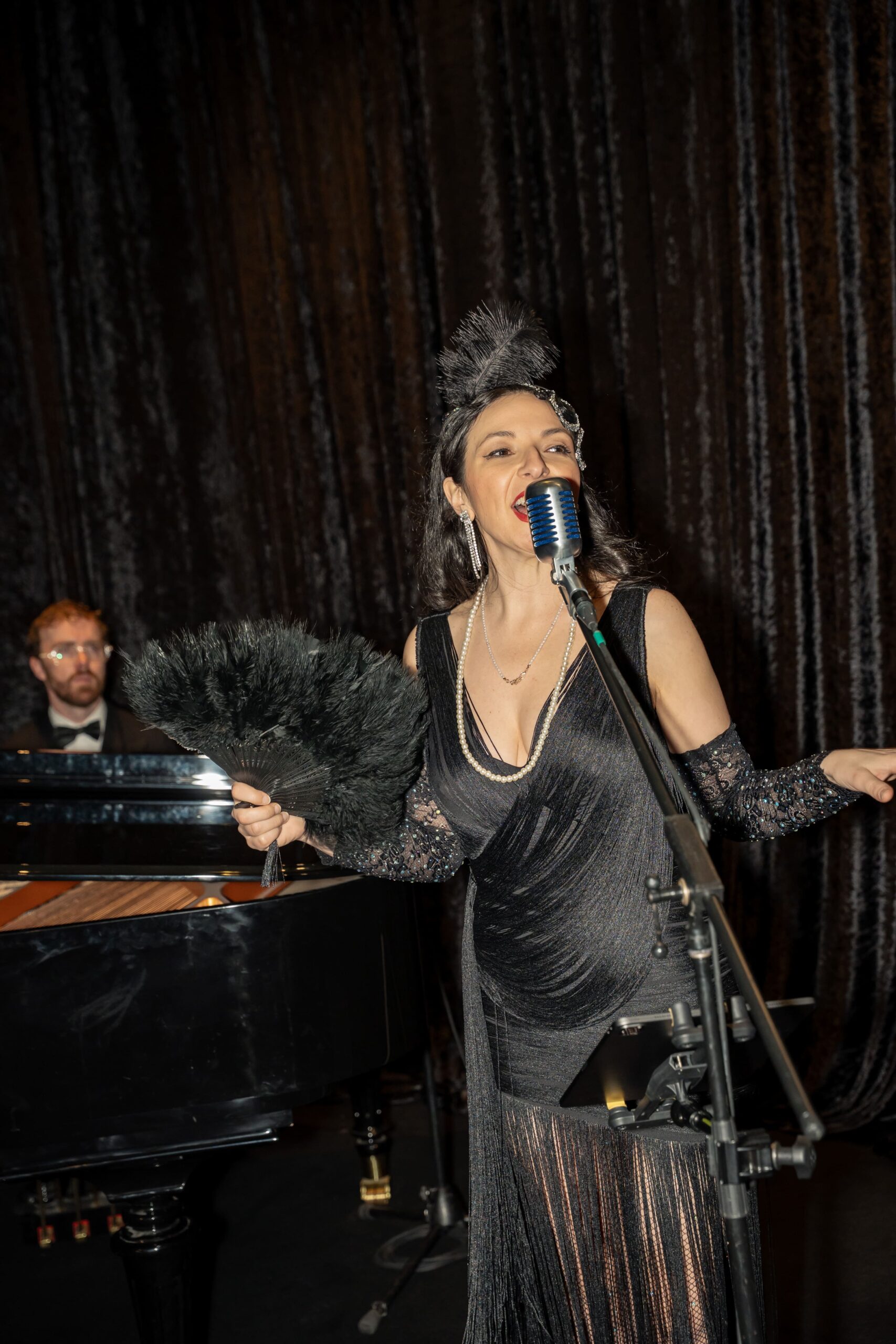 A woman in a black flapper-style dress sings into a vintage microphone, holding a feather fan, while a pianist in a tuxedo plays a grand piano in the background against dark curtains at Million Dollar Lunch 2025