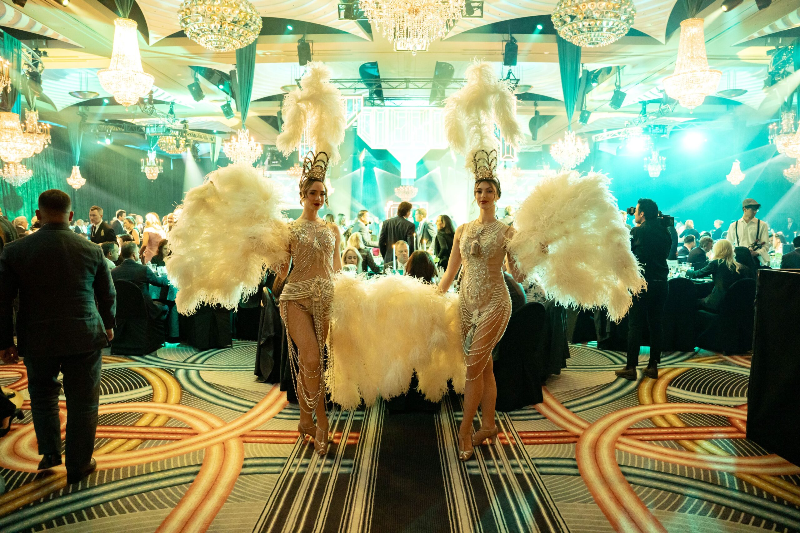 Two showgirls in sparkling costumes and feathered headdresses stand in a glamorous, chandelier-lit ballroom at Million Dollar Lunch 2025, where guests seated at tables enjoy a luxurious and festive atmosphere.